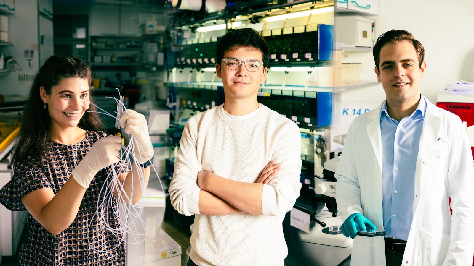 Three scientists standing inside a laboratory. The female scientist on the left is holding what looks like a set of narrow tubing. 