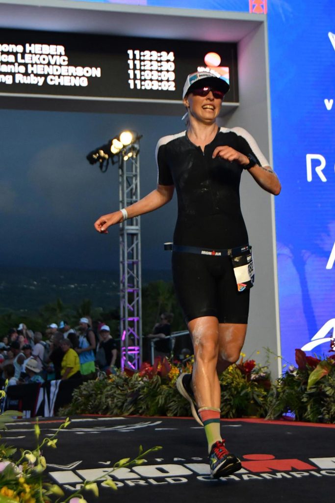 Woman runner on a race track in the foreground, with audiences visible in the background. A scoreboard on the top left shows the timings of various participants in the triathlon.