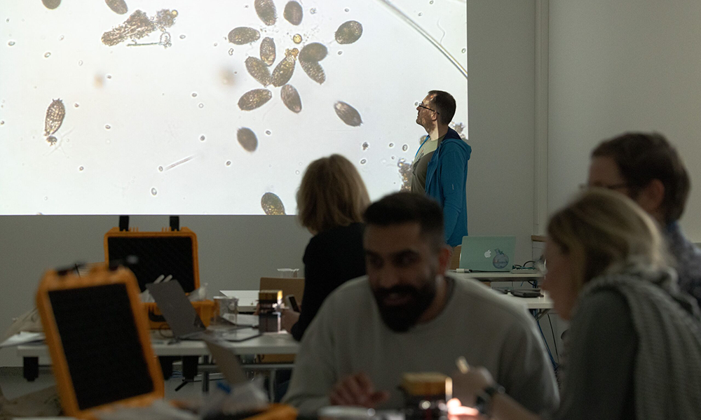 A man is standing in front of a slide showing a group of microorganisms. Several participants in front of the slide are sitting at tables equipped with Curiosity microscopes.