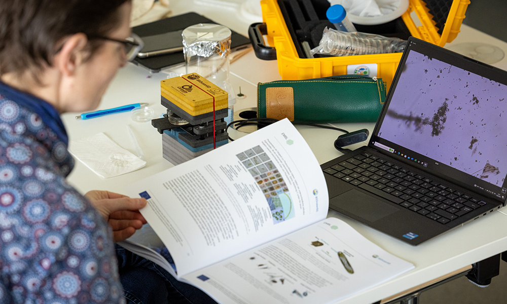 A woman is looking through a book and checking a microscopy image on the screen of a laptop in front of her. On her left, there is the Curiosity microscope, which consists of grey, blue and yellow blocks connected with a red stripe.