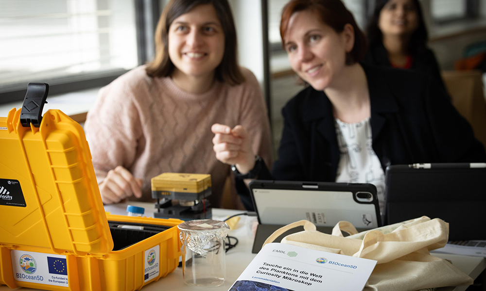Two smiling women are looking and pointing at something outside of the picture. In front of them, there is a Curiosity microscope, a laptop and an EMBL guide booklet.