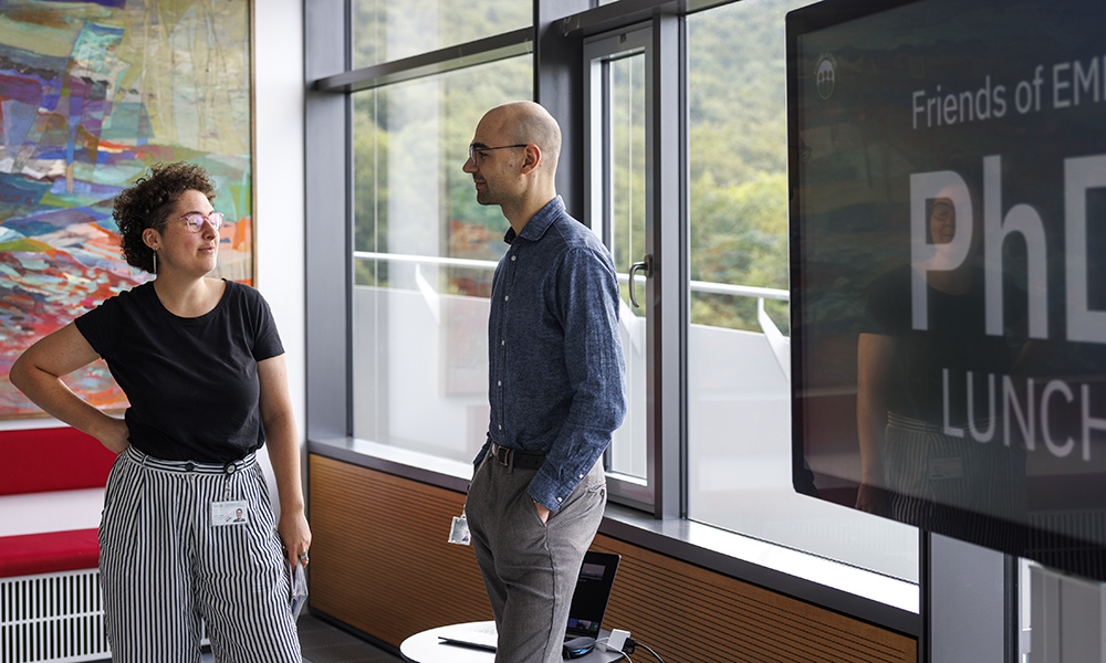 Two scientists stand in front of window, talking