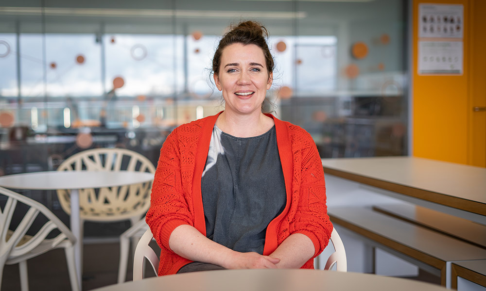 Woman wearing grey top and orange cardigan sat at a table, smiling.
