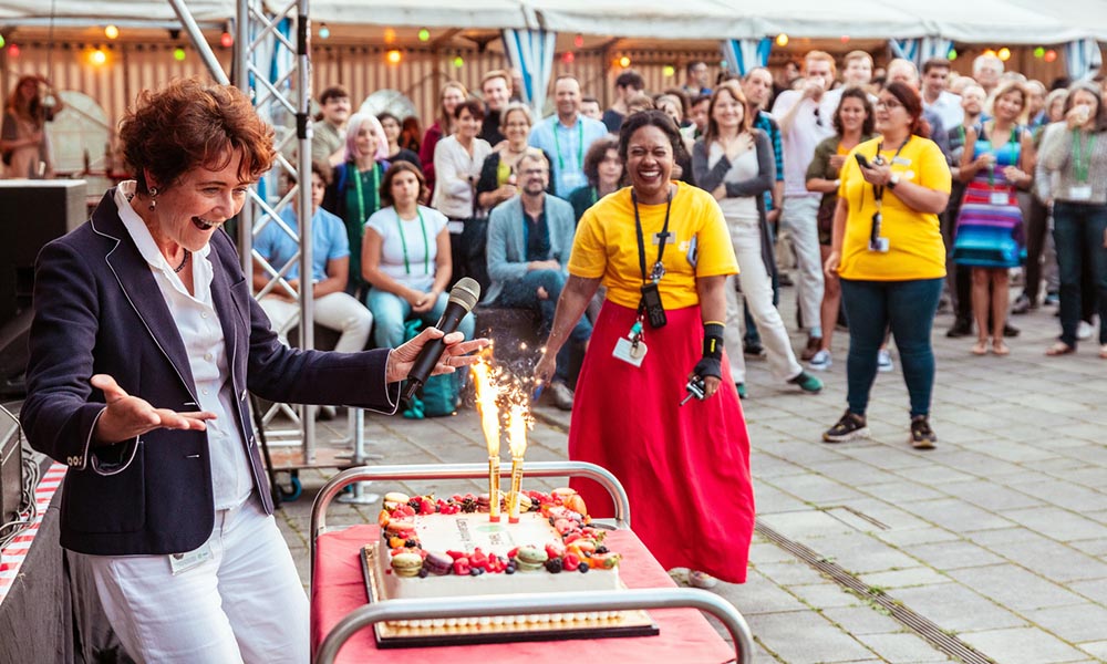 Director General stands in front of a crowd with a celebratory anniversary cake alight with sparklers.
