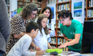 Image showing three adults and two children inside a library. One of the adults is an EMBL volunteer and has her finger inside a green bowl next to a pair of tongs. The children are looking inside the bowl.