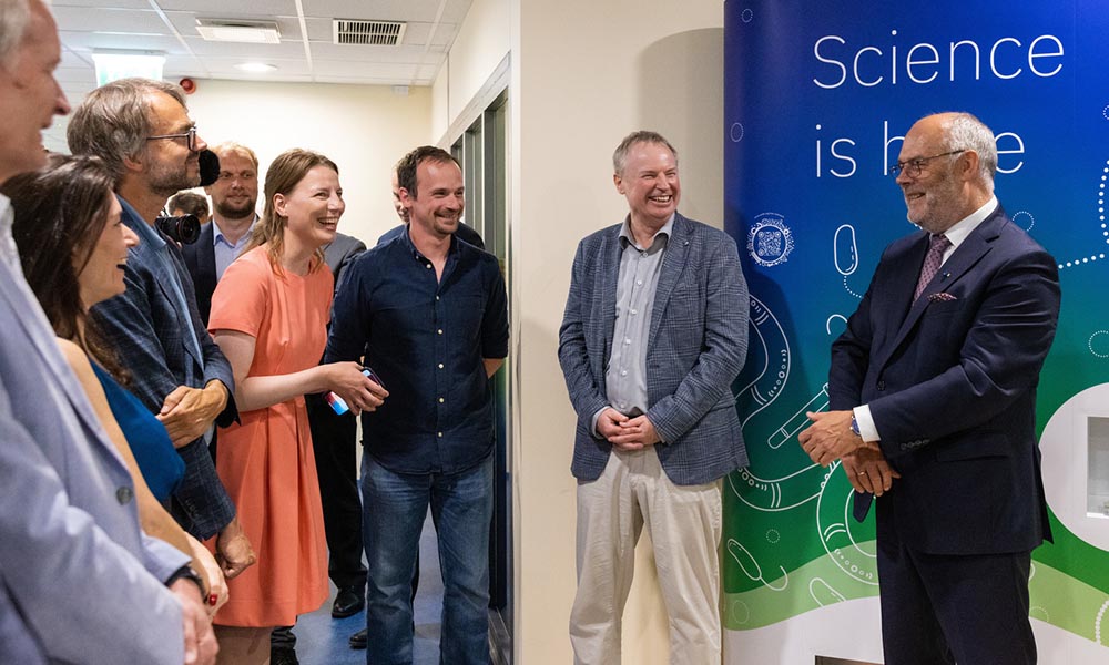 Estonian president stands in front of blue poster, talking to group of scientists