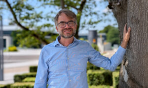Male scientist in blue shirt standing next to a tree outdoors.