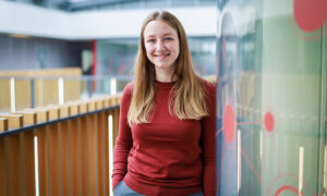 Young woman with long, blond hair, wearing a dark red top, leaning against a glass wall and smiling. Credit: Jeff Dowling