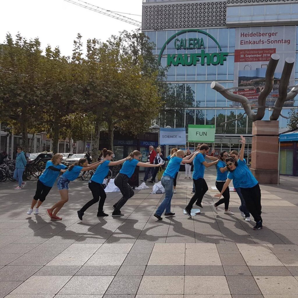 Scientists wearing blue t-shirts perform hip-hop choreography in a public venue in Heidelberg.
