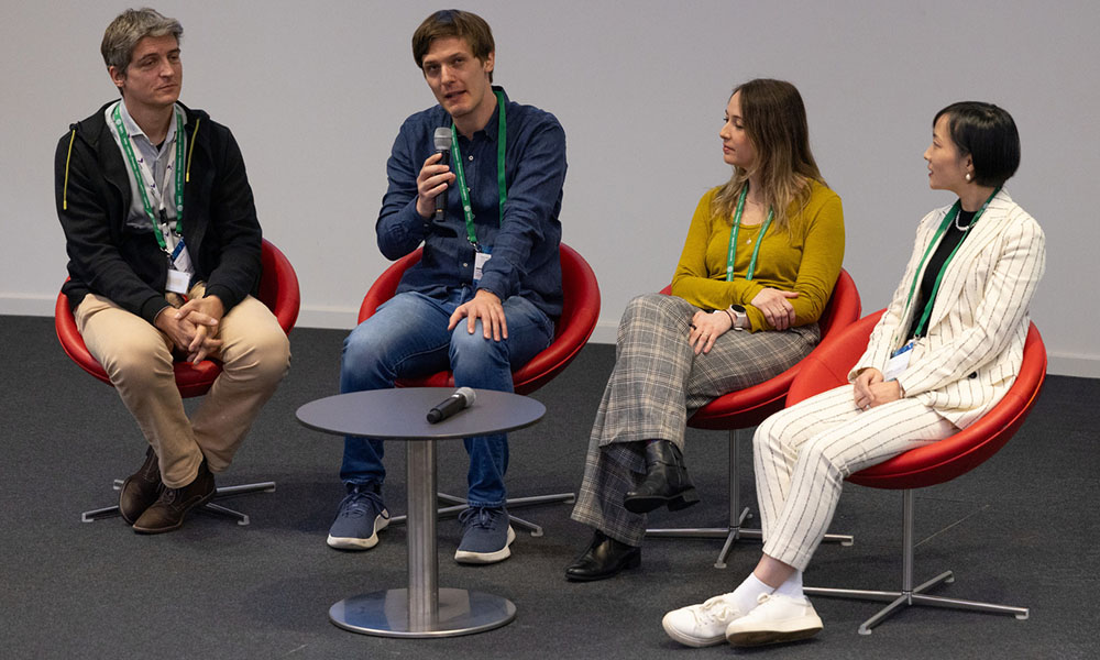 Four panelists sit onstage, facing toward the camera and sharing a handheld microphone.