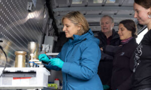 Rhineland-Palatinate Climate Protection Minister Katrin Eder in blue jacket, tries out the sampling process in one of TREC's mobile labs. EMBL colleagues stand next to her.