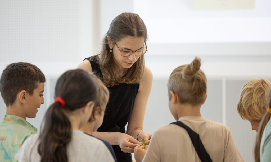 A woman demonstrating something to a group of four children with a leaf in her hand.