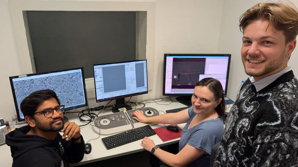 Three scientists in front of monitors associated with microscopes.