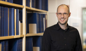 A man in a black shirt in front of a bookshelf containing several theses bound in blue.