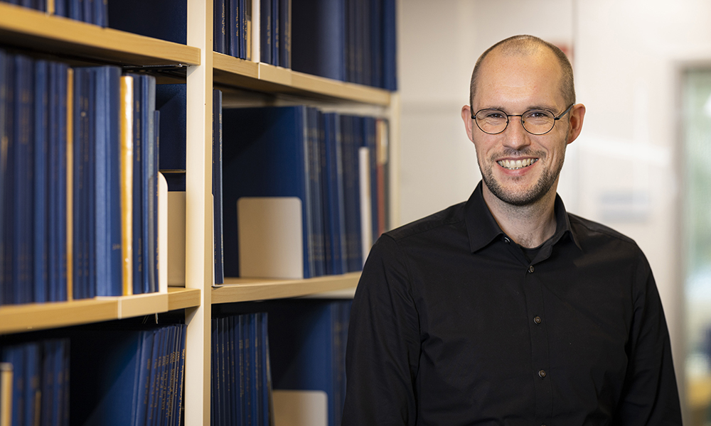 A man in a black shirt in front of a bookshelf containing several theses bound in blue.