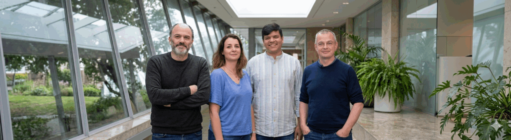 Four adults stand side by side in a bright, modern indoor hallway with large glass windows and greenery visible outside. The group faces the camera, smiling or with relaxed expressions, dressed casually.