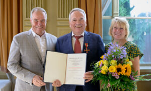 Two men and a woman posing for a photograph. The man in the centre is wearing the German Federal Cross and holding a certificate. The woman is holding a bouquet of flowers.