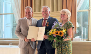 Two men and a woman posing for a photograph. The man in the centre is wearing the German Federal Cross and holding a certificate. The woman is holding a bouquet of flowers.
