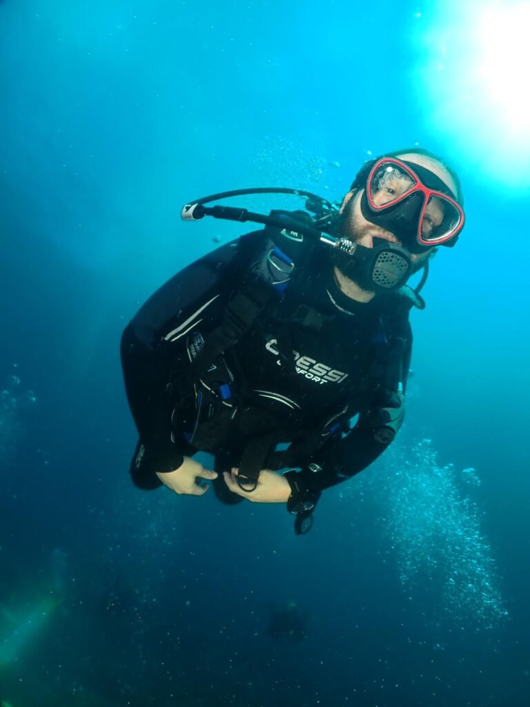 Daniel scuba diving, wearing a black wetsuit, red-and-black diving mask, and breathing apparatus floats underwater in clear blue water, with streams of bubbles rising around him.