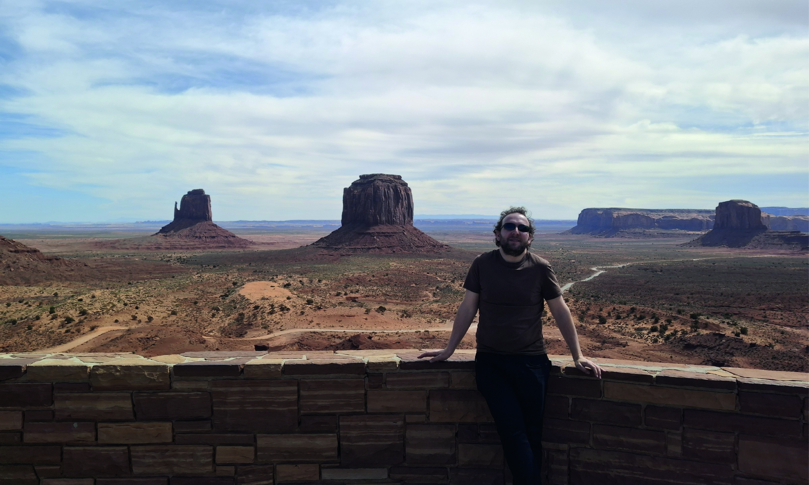 Daniel wearing sunglasses leans against a stone wall at a scenic overlook in Monument Valley, with towering red sandstone buttes and a vast desert landscape beneath a partly cloudy sky.