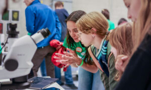Photograph showing two young women looking into a microscope as a volunteer explains how it works.